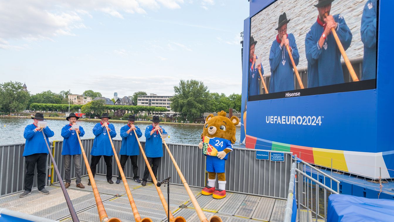 Fünf Herren spielen in landestypischen blauen Hemden Alphorn um das Schweizer Fußball-Team anzufeuern. 