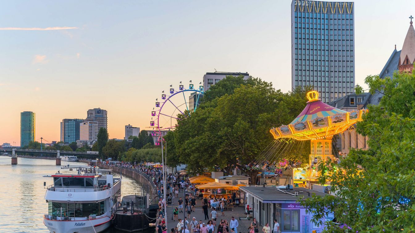 Blick auf ein beleuchtetes Karussell und ein Riesenrad am Frankfurter Mainufer bei Sonnenuntergang, mit Menschen, die an den Ständen entlang spazieren.