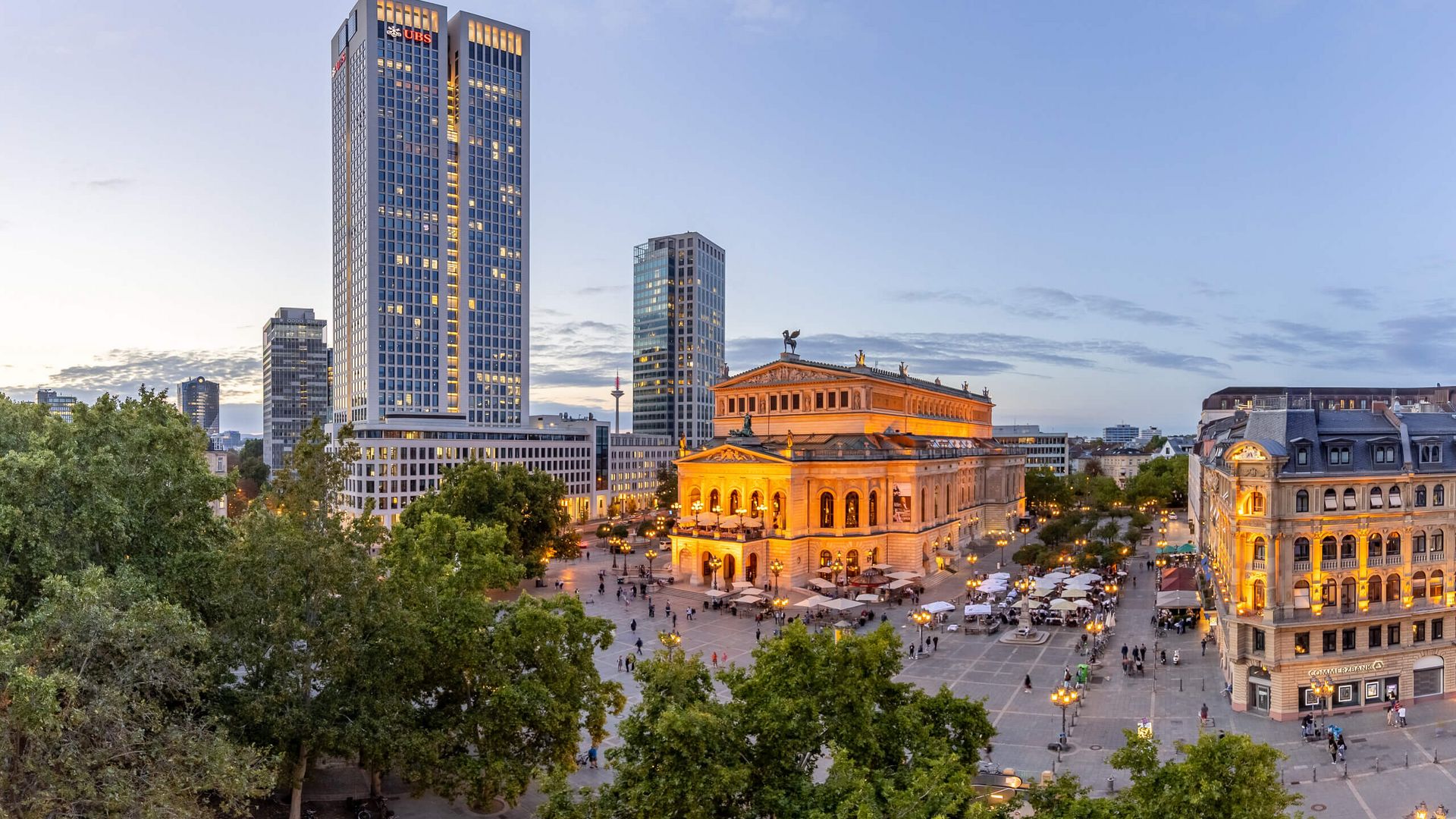 Panoramabild mit Alter Oper, Hochhaus und schönen Altbauten, Laternen tauchen den Platz in gelbes Licht, Abendstimmung.