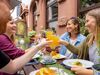 Eine Gruppe Menschen stößt mit Apfelweingläsern an, vor ihnen auf dem Tisch steht traditionelles Frankfurter Essen. 