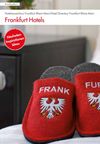 Das Bild zeigt ein Paar rote Hausschuhe mit dem Schriftzug „FRANK“ und „FURT“ auf den jeweiligen Schuhen. Auf den Hausschuhen ist zudem ein Wappen abgebildet. Sie liegen auf einem Teppich in einem Hotelzimmer, mit einem Nachttisch im Hintergrund.