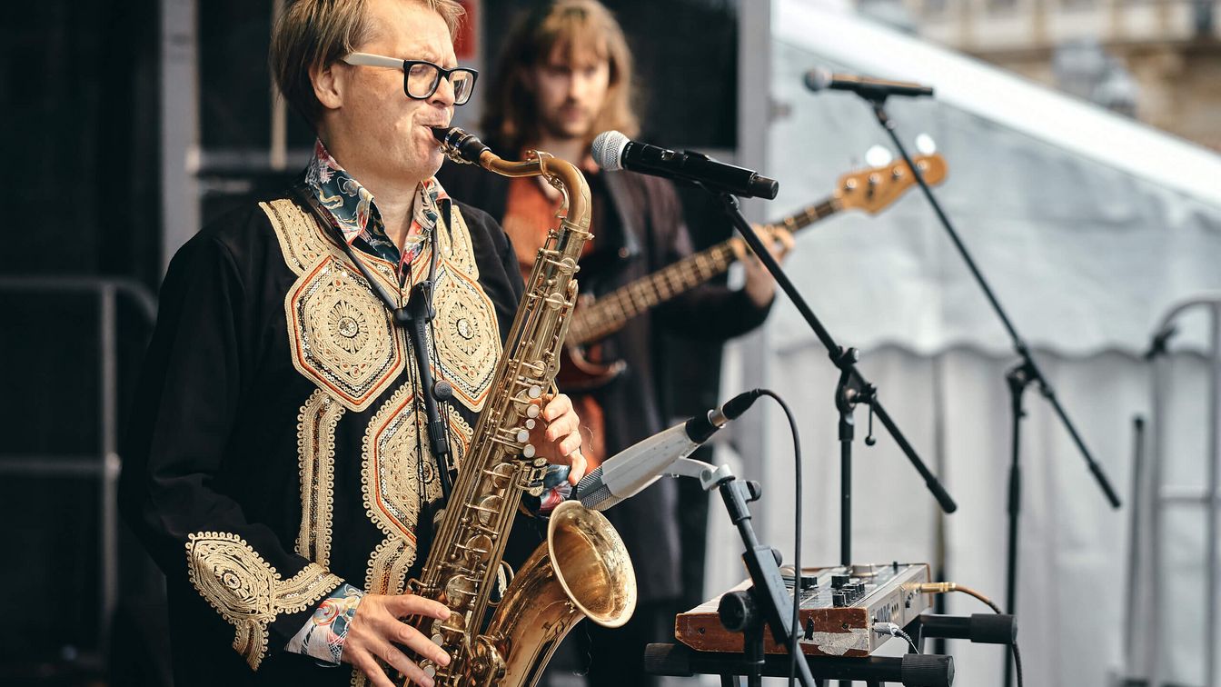 Ein Saxophonist spielt auf einer Bühne, begleitet von einem Bassisten. Mikrofone und Musikinstrumente umgeben sie.