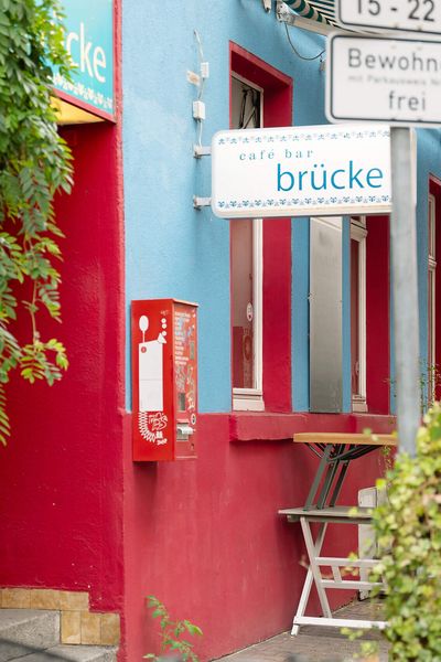 Die Café-Bar „Brücke“ mit leuchtend blauer und roter Fassade, einladender Terrasse und grünen Pflanzen, ergänzt durch orangefarbene Wegweiser im Vordergrund.
