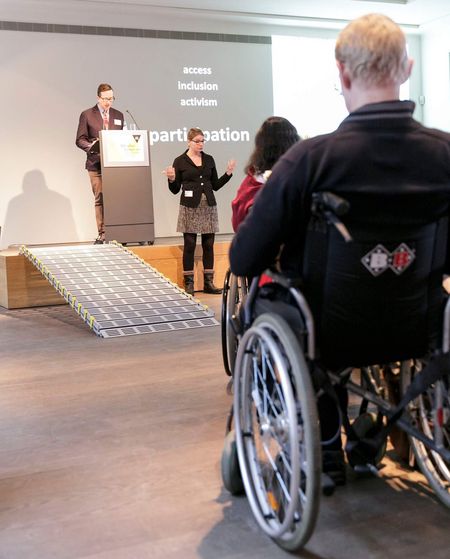 Ein Mann im Rollstuhl lauscht dem Vortrag eines Redners. Der Vortrag wird in Gebärdensprache übersetzt.