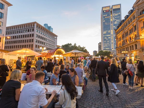 Das Abendlicht beleuchtet den Rheingauer Weinmarkt. Besucher*innen schlendern zwischen Ständen mit Sonnenschirmen und Lichterketten. Die Skyline und historische Gebäude schaffen eine besondere Atmosphäre.