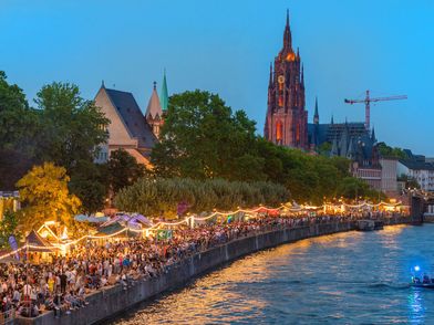 Am Ufer des Mains feiert Menschen am Abend das Museumsuferfest. Der Dom im Hintergrund ist beleuchtet. 