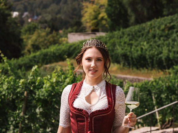 Frau mit Krone im Dirndl genießt Wein im Weinberg vor Stadtkulisse.