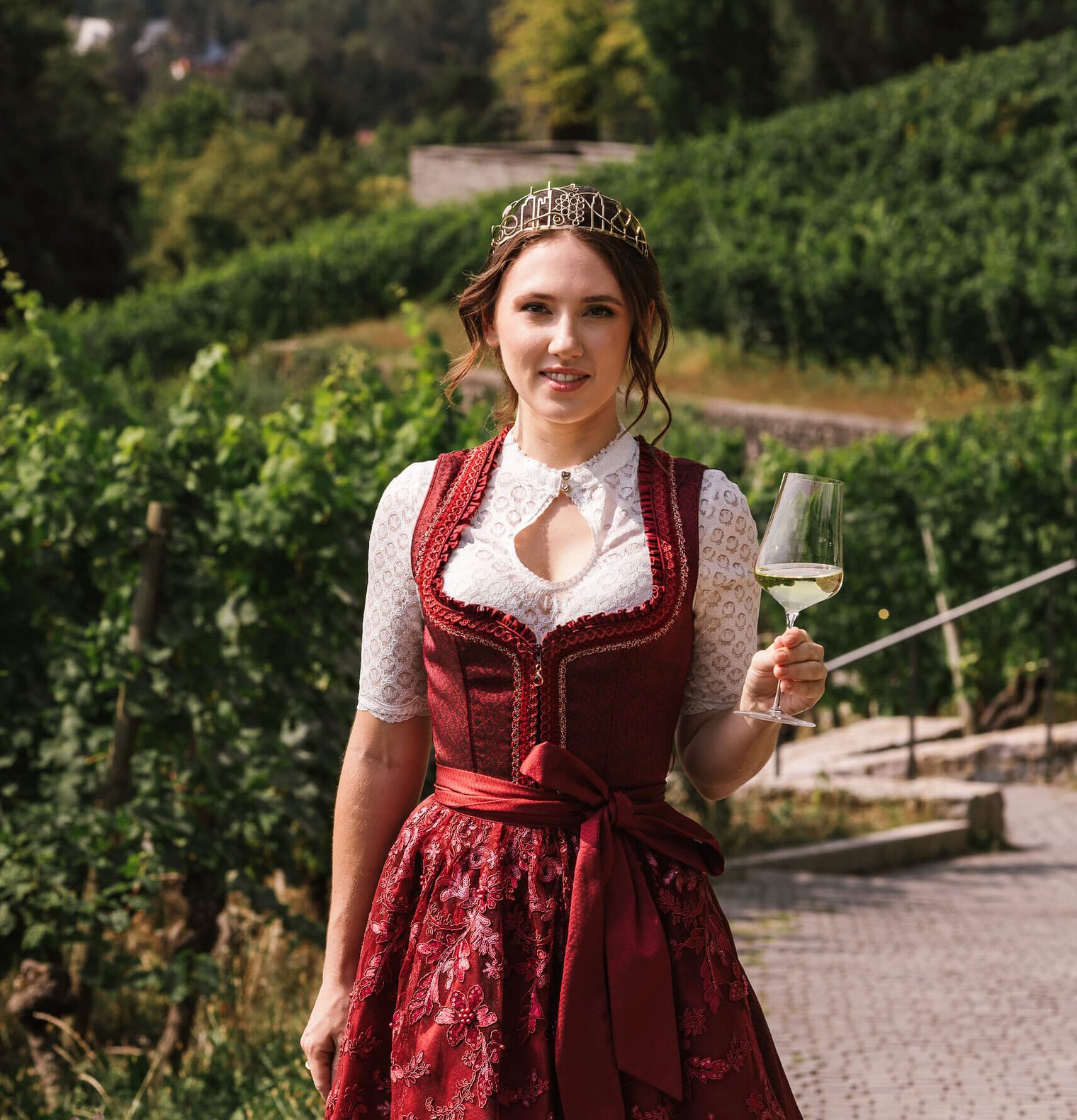 Woman in dirndl enjoying wine in vineyard with city skyline backdrop.