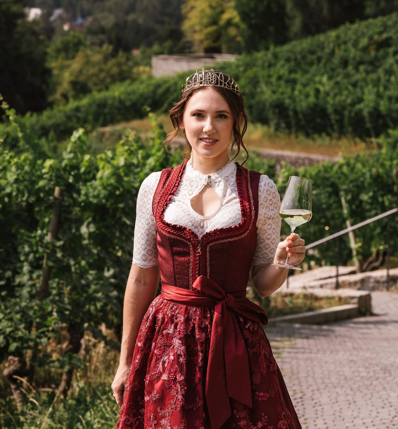 Frau mit Krone im Dirndl genießt Wein im Weinberg vor Stadtkulisse.