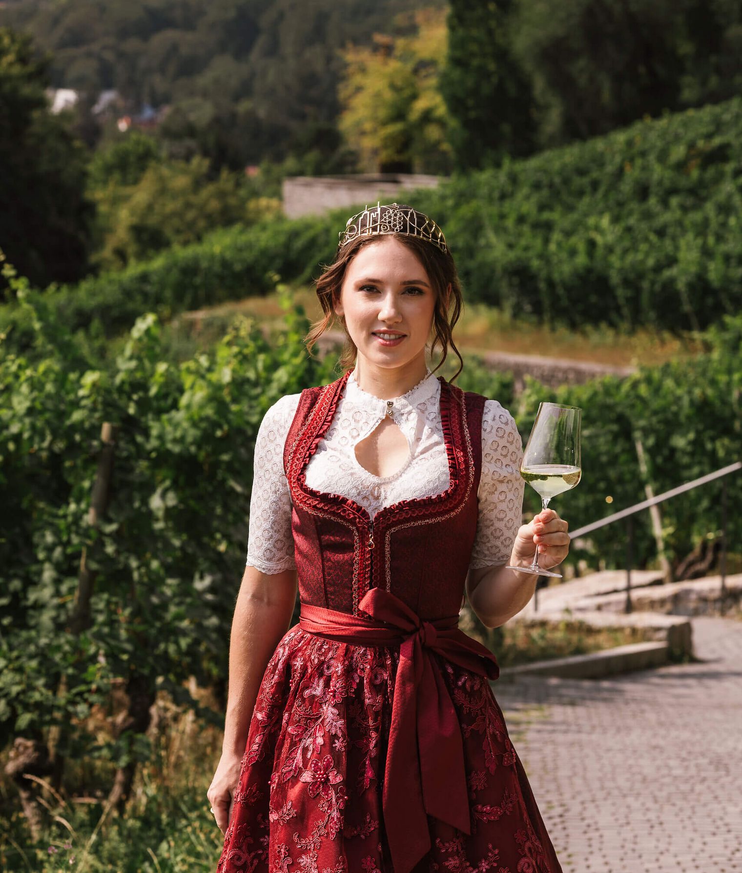 Frau mit Krone im Dirndl genießt Wein im Weinberg vor Stadtkulisse.