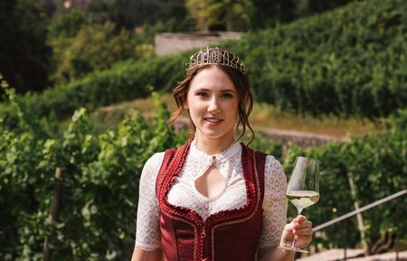 Woman in dirndl enjoying wine in vineyard with city skyline backdrop.