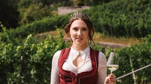 Frau mit Krone im Dirndl genießt Wein im Weinberg vor Stadtkulisse.