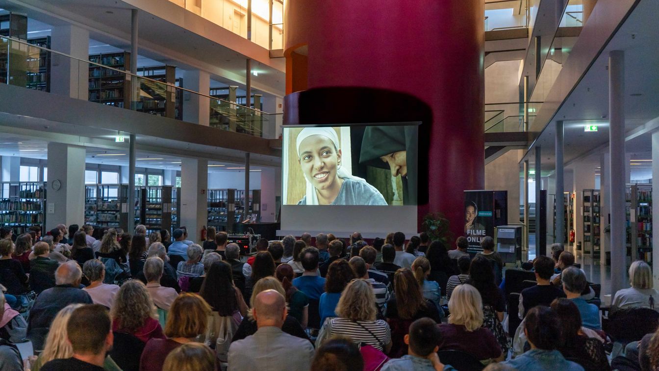 Publikum verfolgt eine Filmvorführung in einer modernen Bibliothek mit großer Leinwand.