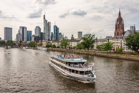 Ausflugsschiff mit Menschen fährt auf dem Fluss Main mit der Frankfurter Skyline und dem Kaiserdom im Hintergrund