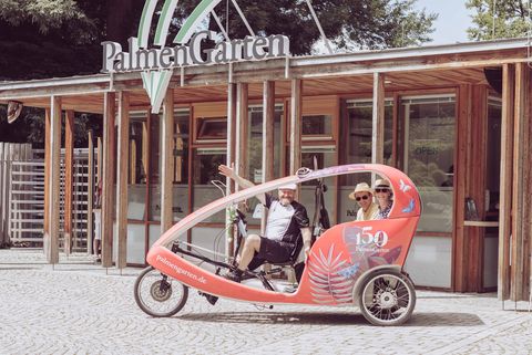Velotaxi vor dem Eingang des Palmengartens in Frankfurt mit zwei lachenden Fahrgästen.