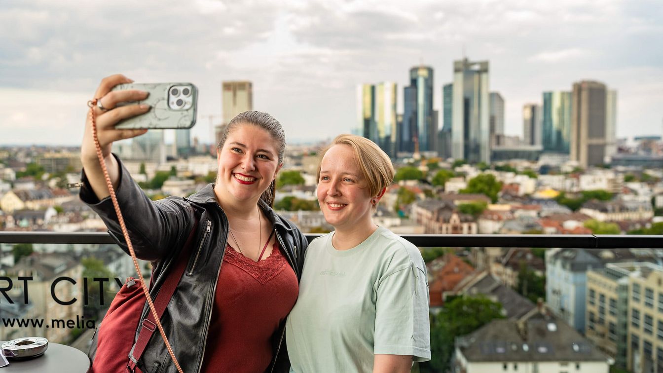 Zwei Frauen machen ein Selfie mit Stadtblick im Hintergrund.