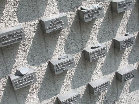 Eine Mauer mit Tafeln, die die Namen deportierter und ermordeter jüdischer Menschen tragen, jeweils mit Gedenksteinen versehen.