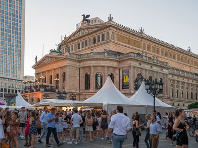 Lebendiges Stadtfest vor der Alten Oper in Frankfurt. Menschen genießen das sommerliche Ambiente mit weißen Festzelten, während moderne Hochhäuser den historischen Bau umrahmen.