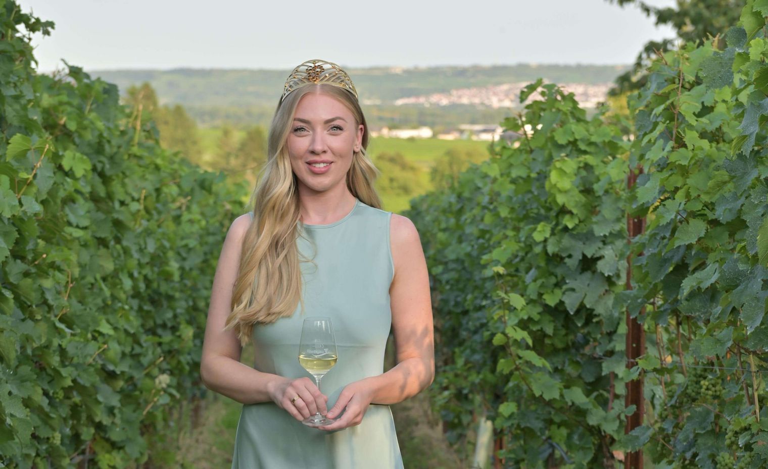 Wine queen wearing a dress and crown in a vineyard with a glass of white wine.