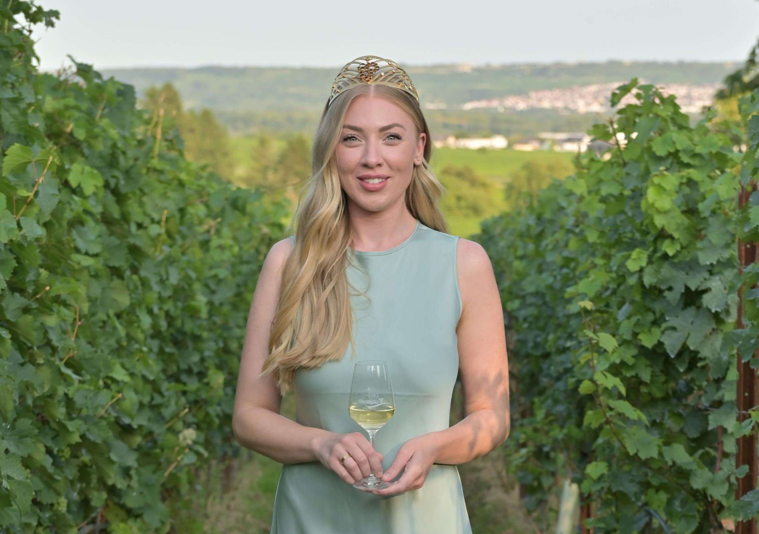 Weinkönigin mit Kleid und Krone im Weinberg mit einem Glas Weißwein.