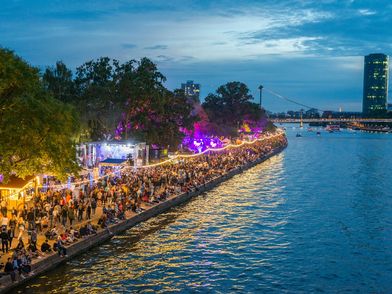 Abendliches Museumsuferfest am Mainufer in Frankfurt mit bunten Lichtern, feiernden Menschen und der beeindruckenden Skyline im Hintergrund.
