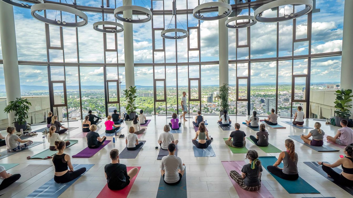Yoga-Gruppe meditiert in einem lichtdurchfluteten Hochhausraum mit weitem Blick über Frankfurt.