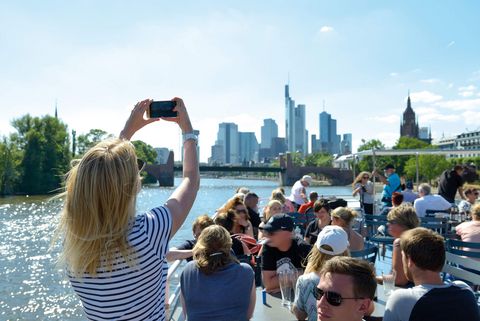 Frau auf dem Sonnendeck eines Schiffes auf dem Main, die ein Foto von der Frankfurter Skyline im Hintergrund macht und weitere Menschen an Tischen, die die Sonne und Getränke genießen.