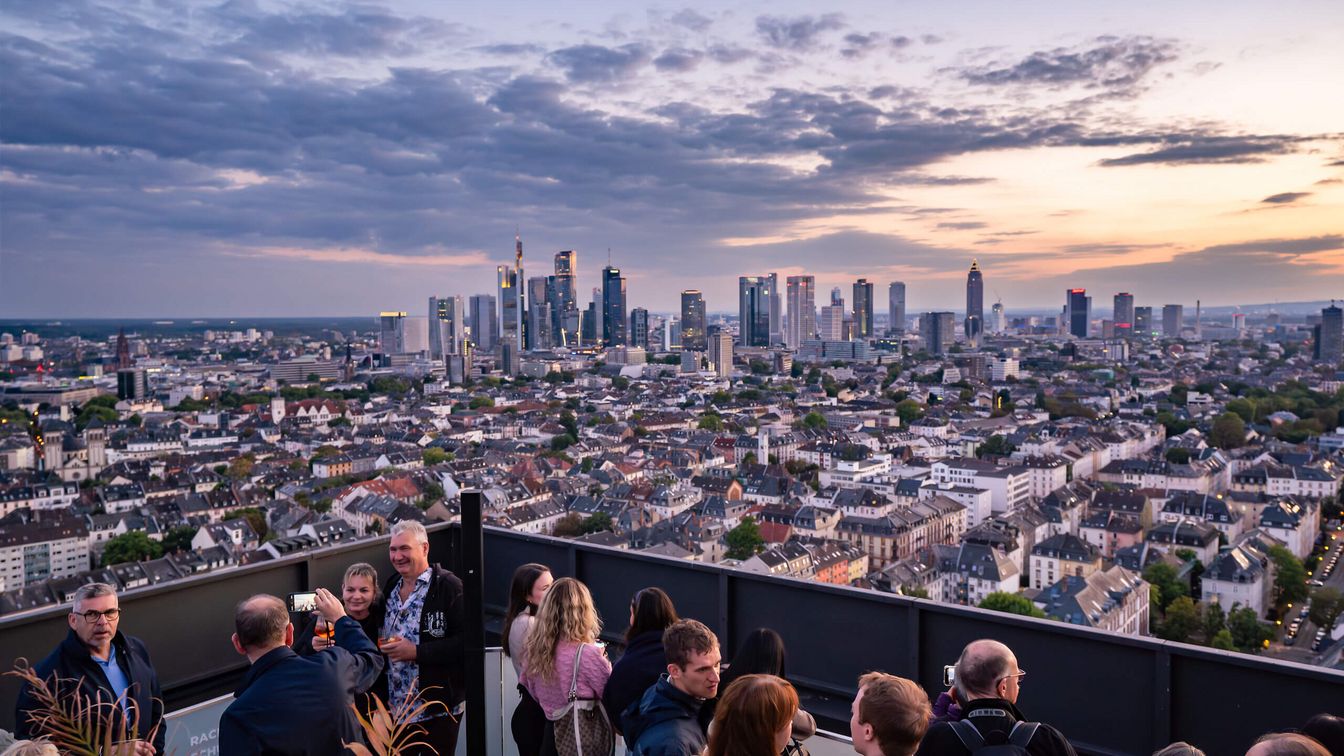 Menschen genießen den Sonnenuntergang auf einer Dachterrasse mit Blick auf die Frankfurter Skyline.