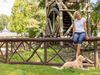 A woman sits on a wooden fence in front of a water wheel, a dog lies at her feet. 