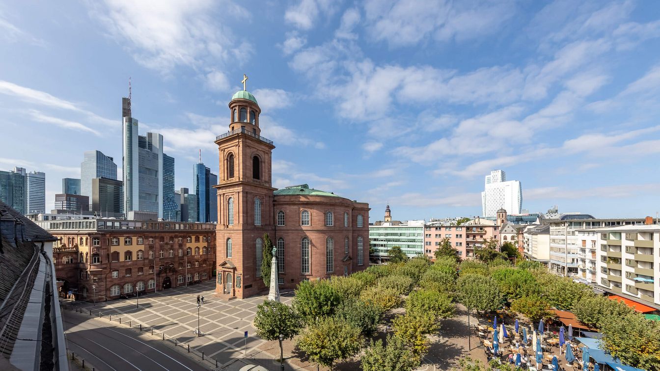 Außenansicht der Frankfurter Paulskirche aus rotem Sandstein, umgeben von Bäumen und einem großen Platz. Im Hintergrund ragen moderne Hochhäuser der Frankfurter Skyline in den Himmel, der von leichten Wolken bedeckt ist.