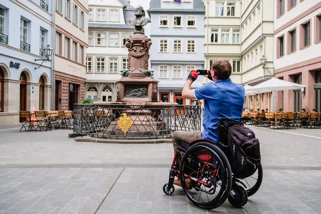 A man in a wheelchair takes a photo of the Stoltze fountain at the Hühnermarkt