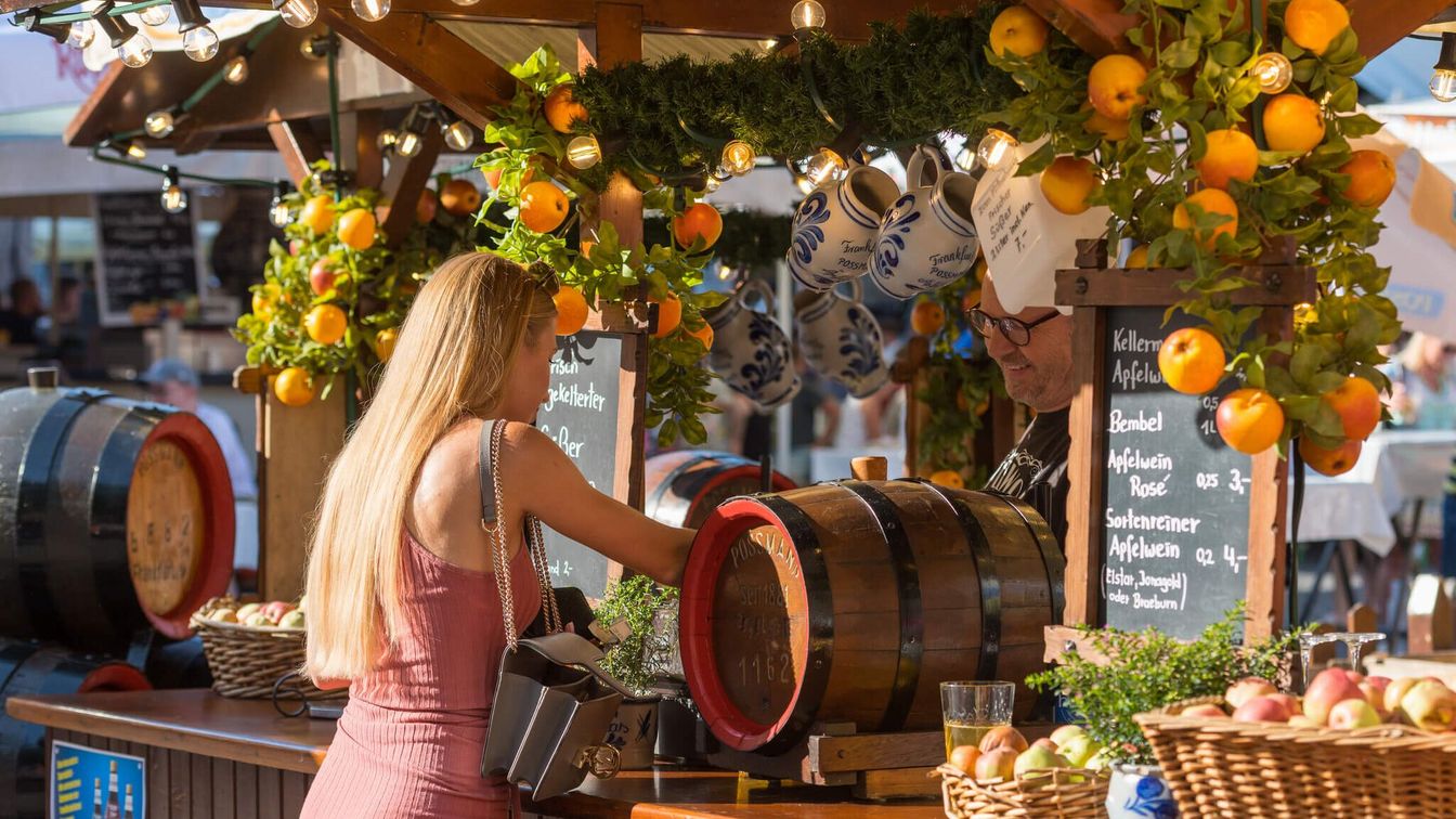 A woman is standing at a cider stall decorated with apples and bembels (earthenware jugs) 