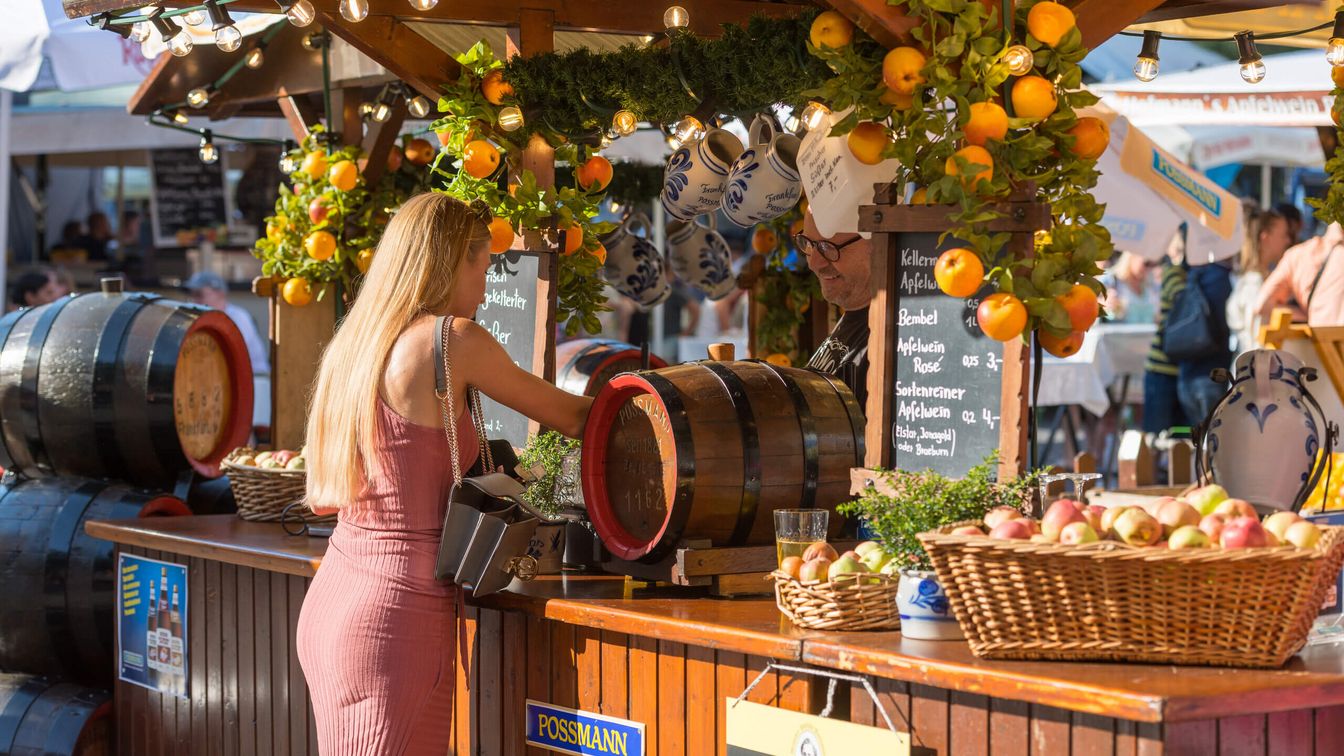A woman is standing at a cider stall decorated with apples and bembels (earthenware jugs) 