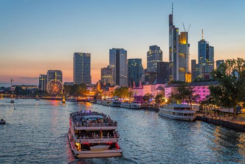 Blick auf den Main, Schiffe der Primus Linie und die dahinterliegende Frankfurter Skyline in der Abenddämmerung, am Ufer leuchtende Stände und ein Riesenrad. 