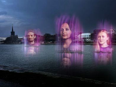 Leuchtende, holographische Gesichter schweben über einem Gewässer in Frankfurt bei Nacht. Die Lichtinstallation erzeugt eine mystische Atmosphäre mit der Skyline im Hintergrund.