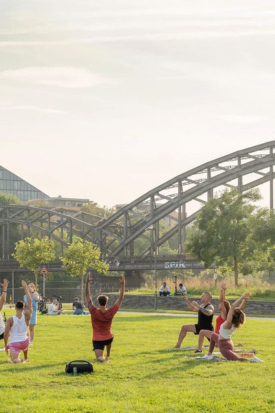 Menschen machen Yoga-Übungen auf einer Wiese, im Hintergrund eine Brücke und ein modernes Gebäude.