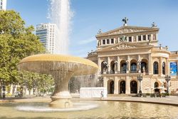 The Alte Oper Frankfurt in bright sunshine, the bubbling Lucae Fountain in the foreground with trees and modern architecture in the background.
