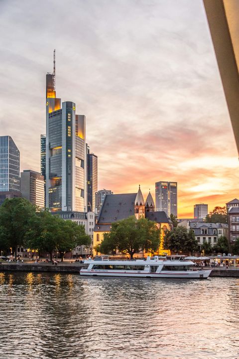 A passenger ship lies in the evening atmosphere on the shore in front of the Leonardskirche and the Commerzbank skyscraper