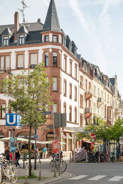 Straßenszene an der Leipziger Straße in Frankfurt mit Altbauten, einem U-Bahn-Schild, Fahrrädern und geschäftigem Treiben bei klarem Himmel.