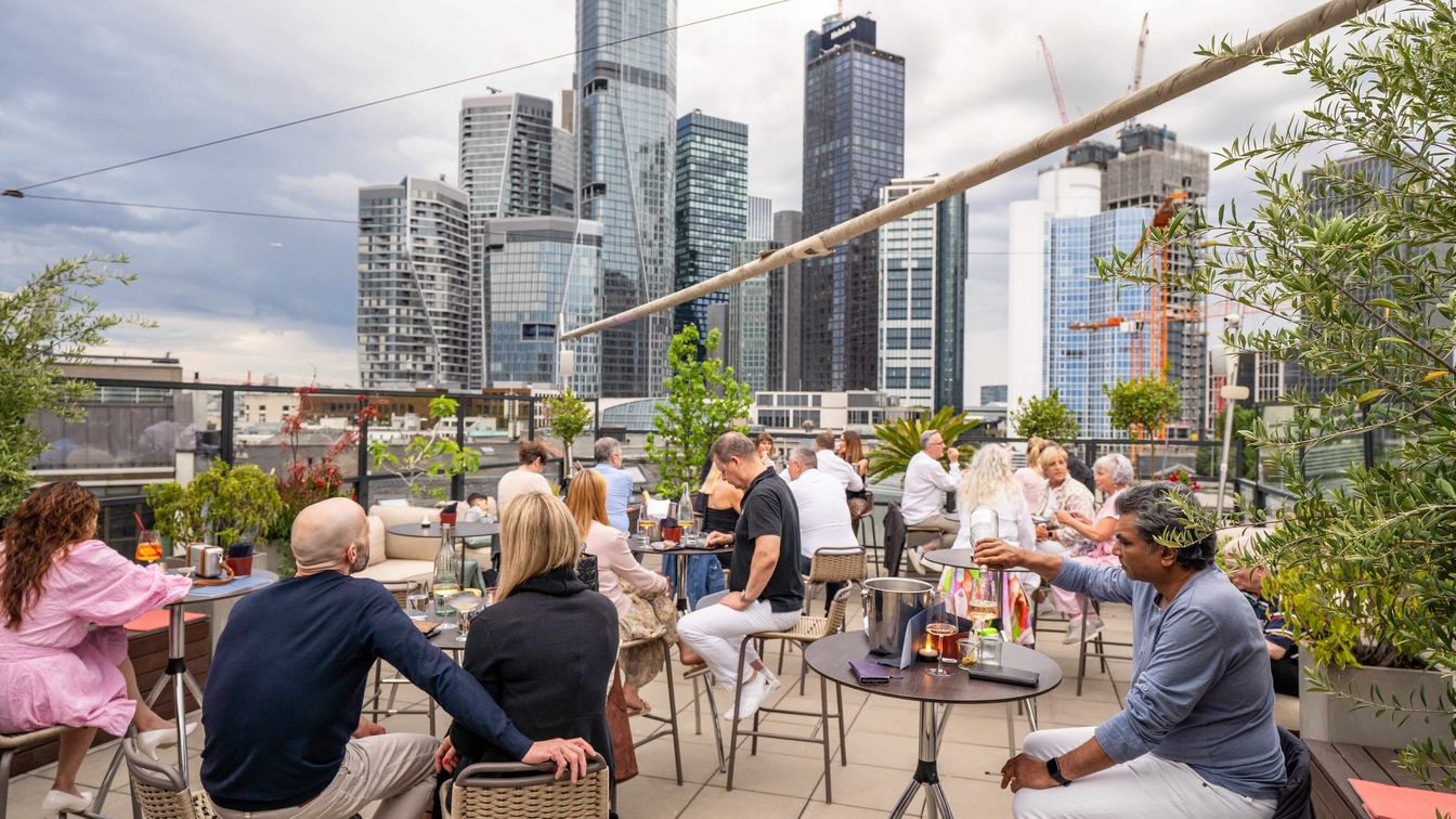 Belebte Dachterrasse mit Pflanzen, Gästen und Blick auf die Frankfurter Skyline unter Wolken.
