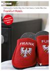 Das Bild zeigt ein Paar rote Hausschuhe mit dem Schriftzug „FRANK“ und „FURT“ auf den jeweiligen Schuhen. Auf den Hausschuhen ist zudem ein Wappen abgebildet. Sie liegen auf einem Teppich in einem Hotelzimmer, mit einem Nachttisch im Hintergrund.