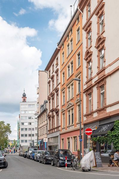 Eine Straßenszene im Frankfurter Brückenviertel mit historischen Gebäuden, Geschäften und Straßencafés. Fahrräder und Autos prägen das lebendige Straßenbild bei klarem Himmel.