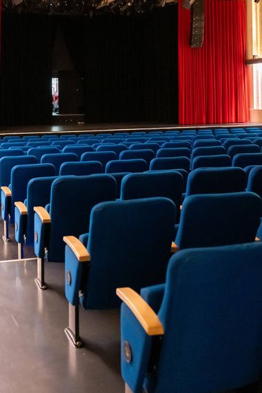 An empty hall with rows of blue velvet-covered seats and a stage with a red curtain. 
