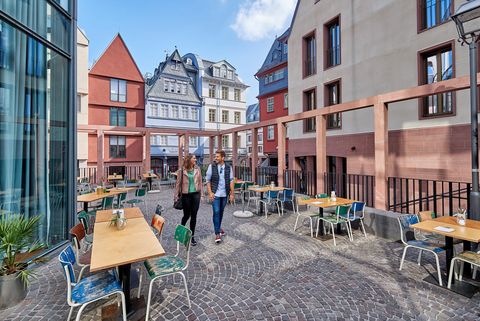 Ein Paar spaziert auf einem gepflasterten Platz in der Frankfurter Altstadt, umgeben von bunten, historischen Gebäuden. Im Vordergrund stehen Tische und Stühle eines Außenbereichs einer Gastronomie.
