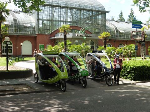 Drei dreirädrigen Velotaxen stehen mit ihren Fahrern und Fahrgästen vor dem Eingang des Palmengartens.