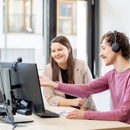 A man and a woman are sitting in front of a computer. The man is explaining something and pointing at the screen.