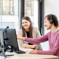 A man and a woman are sitting in front of a computer. The man is explaining something and pointing at the screen.