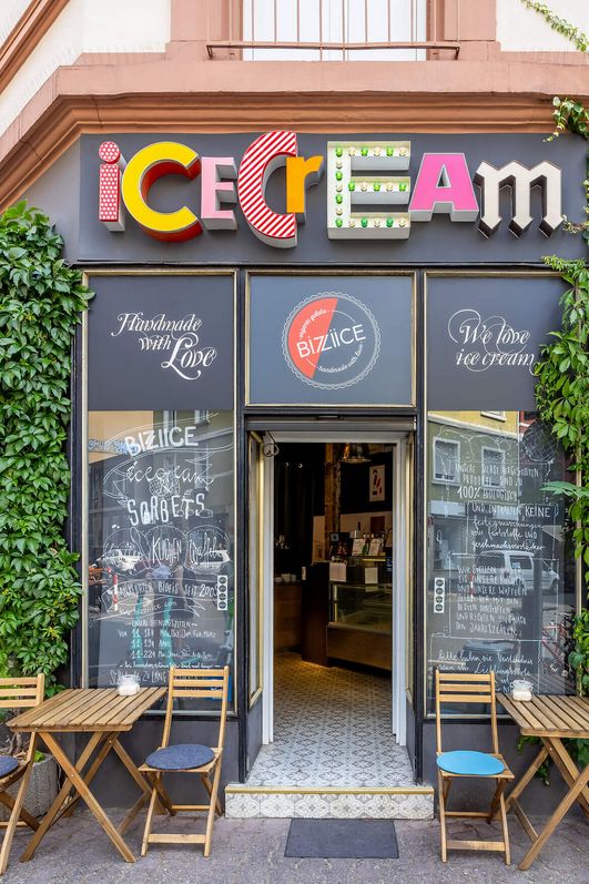 Eisladen mit buntem Schild, Außensitzplatz und Kletterpflanzen im Brückenviertel.
