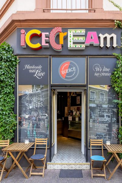Eisladen mit buntem „Ice Cream“-Schild und gemütlicher Außensitzfläche, umgeben von Kletterpflanzen an der Fassade, im Brückenviertel in Frankfurt.