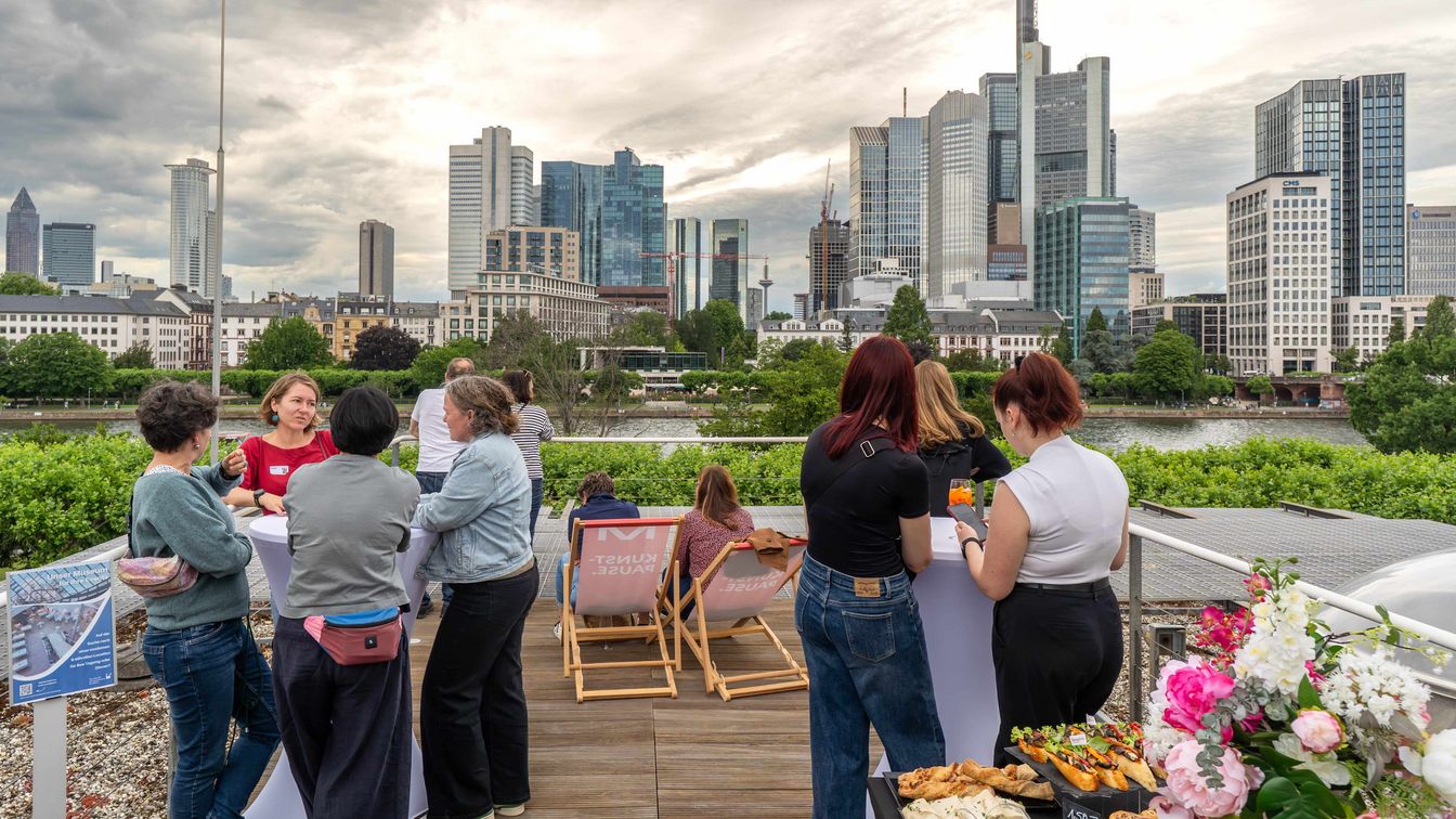 Gäste auf einer Terrasse am Main genießen den Blick auf Frankfurts Skyline und Hochhäuser.
